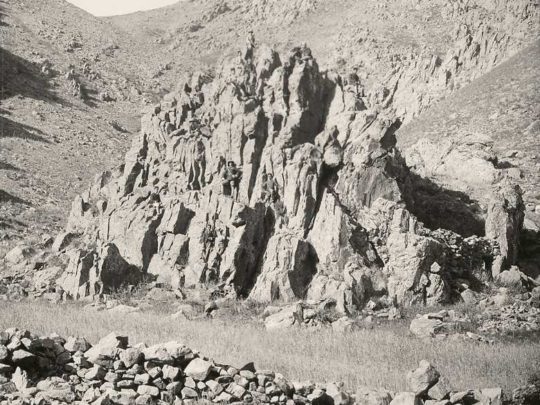 Im Vordergrund ein magerer Acker mit Getreide, von einer Trockenmauer eingefasst; in Bildmitte ein stark zerklüfteter Felsstock auf dem 12 jüngere europäisch gekleidete afghanische Männer stehen oder sitzen.