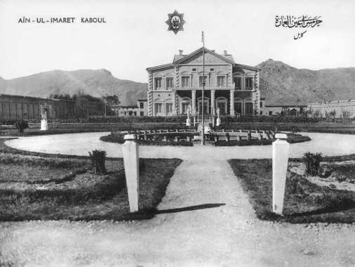 Vue frontale exactement symétrique du palais ayn ul-emarat, et vue de la partie nord-est du jardin, bassin entouré d'un massif de fleurs en pots ; au premier plan deux pilastres blancs. Au centre et devant le bassin, un mât de drapeau ; un réverbère électrique de chaque côté de la maison. Résidence de la reine-mère Uliya Hazrat, construite par le prince Amanullah Khan en 1911-16. Deviendra la légation de l’Iran au début des années 1920. A l’arrière-plan les montagnes asma’i (à droite) et sher darwaza (à gauche). A noter particulièrement la symétrie stricte de la prise et des bâtiments, comme on peut la constater sur d’autres planches de cette série, notamment SdA 2-05 (qawmi bagh), 2-06 et 2-09 (zayn ul-emarat), sur 2-20 (manzel bagh), 2-22 et 2-23 (bagh-e shahi) et sur la planche SdA 2-27 (seraj ul-emarat) ainsi que sur la planche 2-32 (jardin du bagh-e shahi).