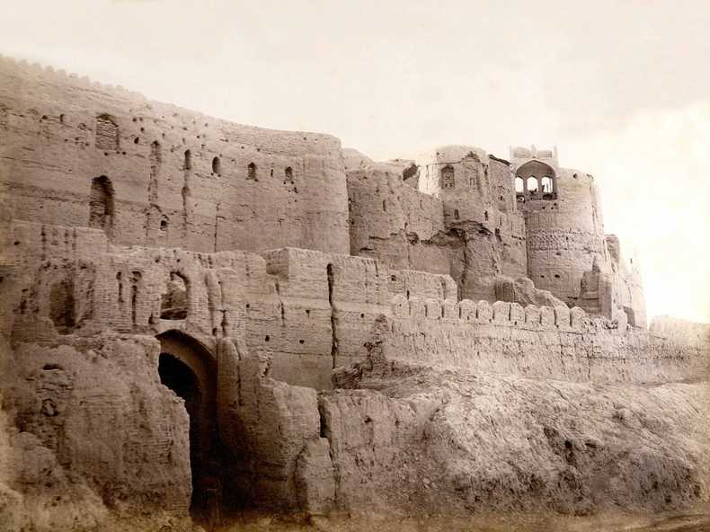 General view of the western front of the citadel with its entrance and the highest remaining parts of the walls. There are three successive walls of increasing height. The outer one with a crenellation, the middle one is rather dilapidated, the over-towering inner walls are partly decorated with geometrical brick patterns and remains of a crenellation. The upper part of the lofty corner tower is fallen in, but some openings with pointed arches remain. In front of the entrance one person in traditional clothing is slightly visible.