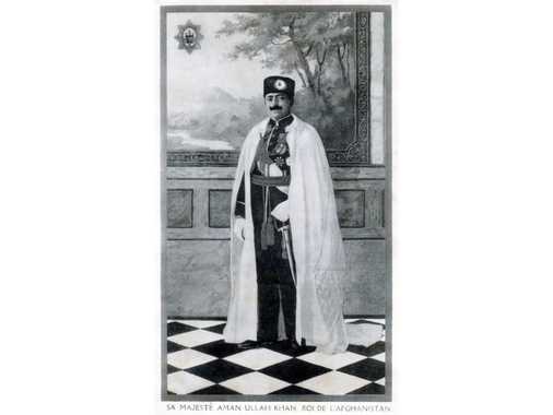 Portrait en pied du roi Amanullah Khan, (1892-1919-1929-1960), fils de l’émir Habibullah Khan (11.1.1.2), né le 1 juillet 1892 à Paghman. Son nom signifie «La Paix Divine» ce qui correspondrait au nom français «Godfrey» ou au nom allemand «Gottfried». En grand uniforme et manteau d’honneur, gants blancs, toque d’astrakhan (kolah) orné d’une décoration. Photo d’atelier, devant un décor de mur de palais imitant un tableau encadré d’un paysage de montagne, une rivière et des arbres.